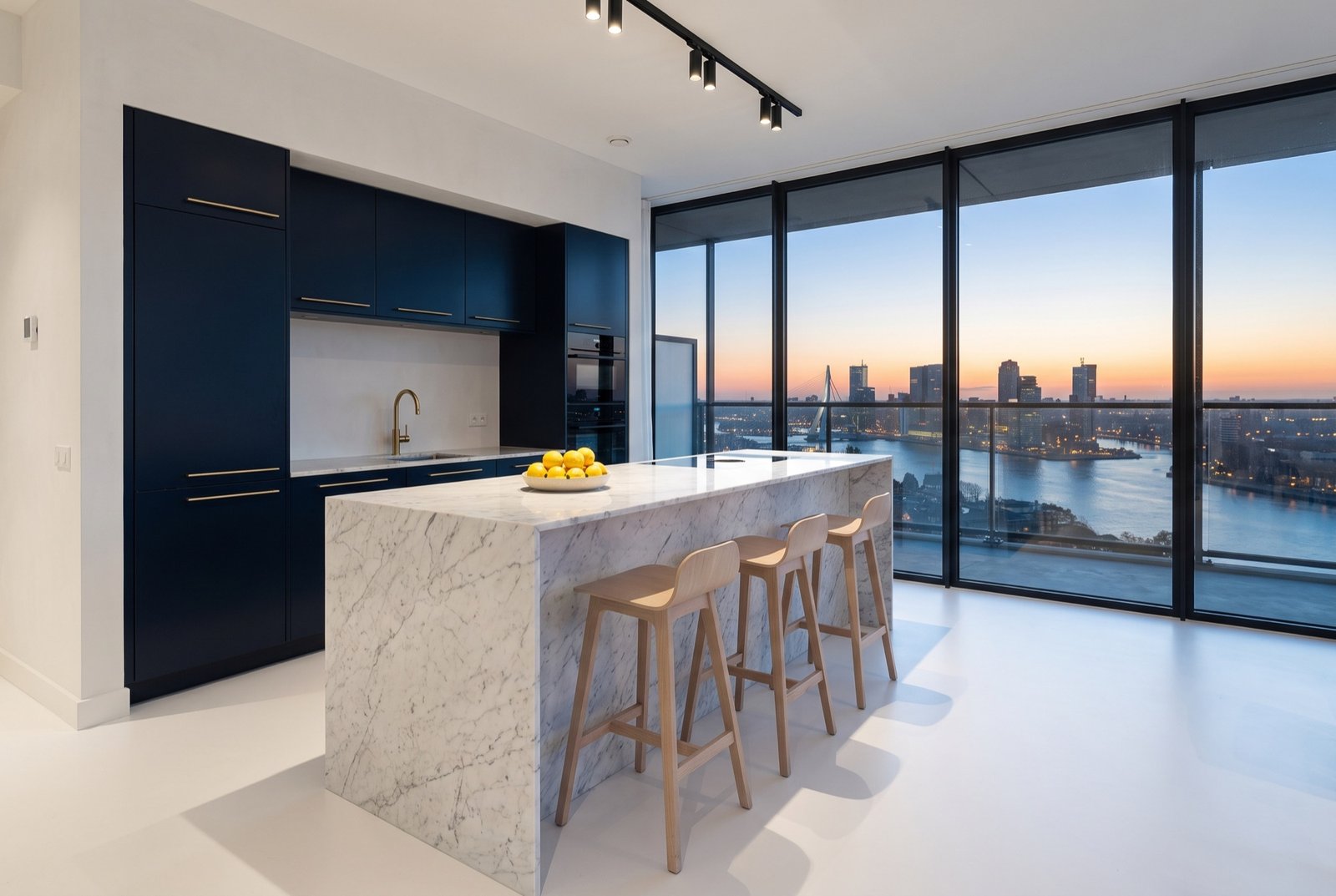 Kitchen with a white marble island, night-blue cabinetry and brass in front of a glass wall at dusk